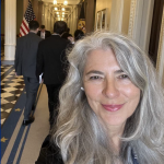 selfie of a women with light skin and silver hair smiling in a hallway that looks like a government building