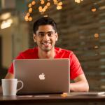 A man in a red shirt and a cup of coffee looks over the the screen of his laptop.