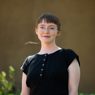 A headshot of Audrie Francis. A person in a black shirt with circular glasses, they have bangs and they are smizing. They have an unfortunate flyaway from their bun hairstyle.