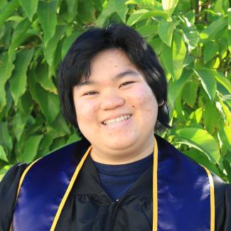 A chest up photo of Shirley. They are wearing their blue and gold Berkeley undergraduate stoll. They are smiling at the camera at a 3/4 face view, pointing to the left. The background consists of bright green tree leaves.