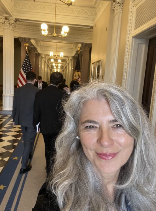 selfie of a women with light skin and silver hair smiling in a hallway that looks like a government building