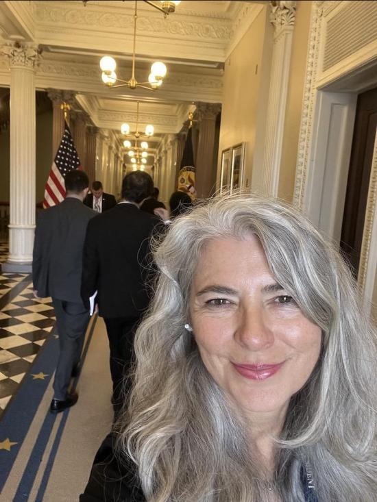 selfie of a women with light skin and silver hair smiling in a hallway that looks like a government building