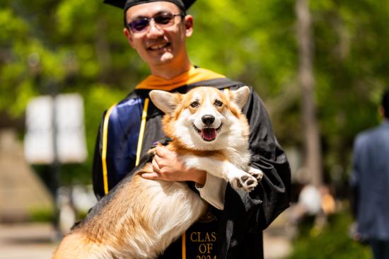 graduate in glasses holds up corgi 