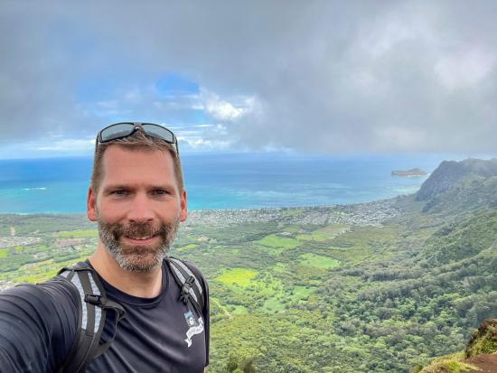 Man in blue shirt and sunglasses smiling at camera with views of the ocean behind him