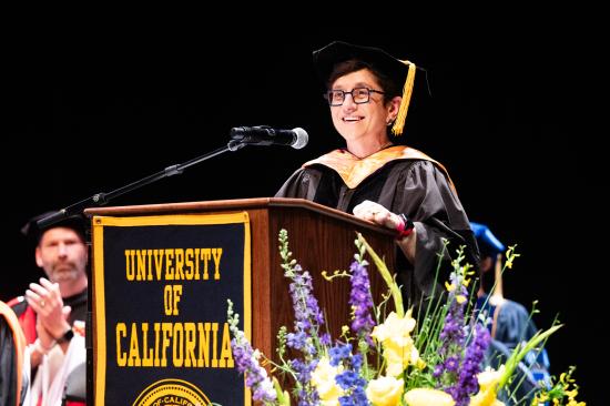 woman with glasses wearing graduation regalia speaking at a lectern